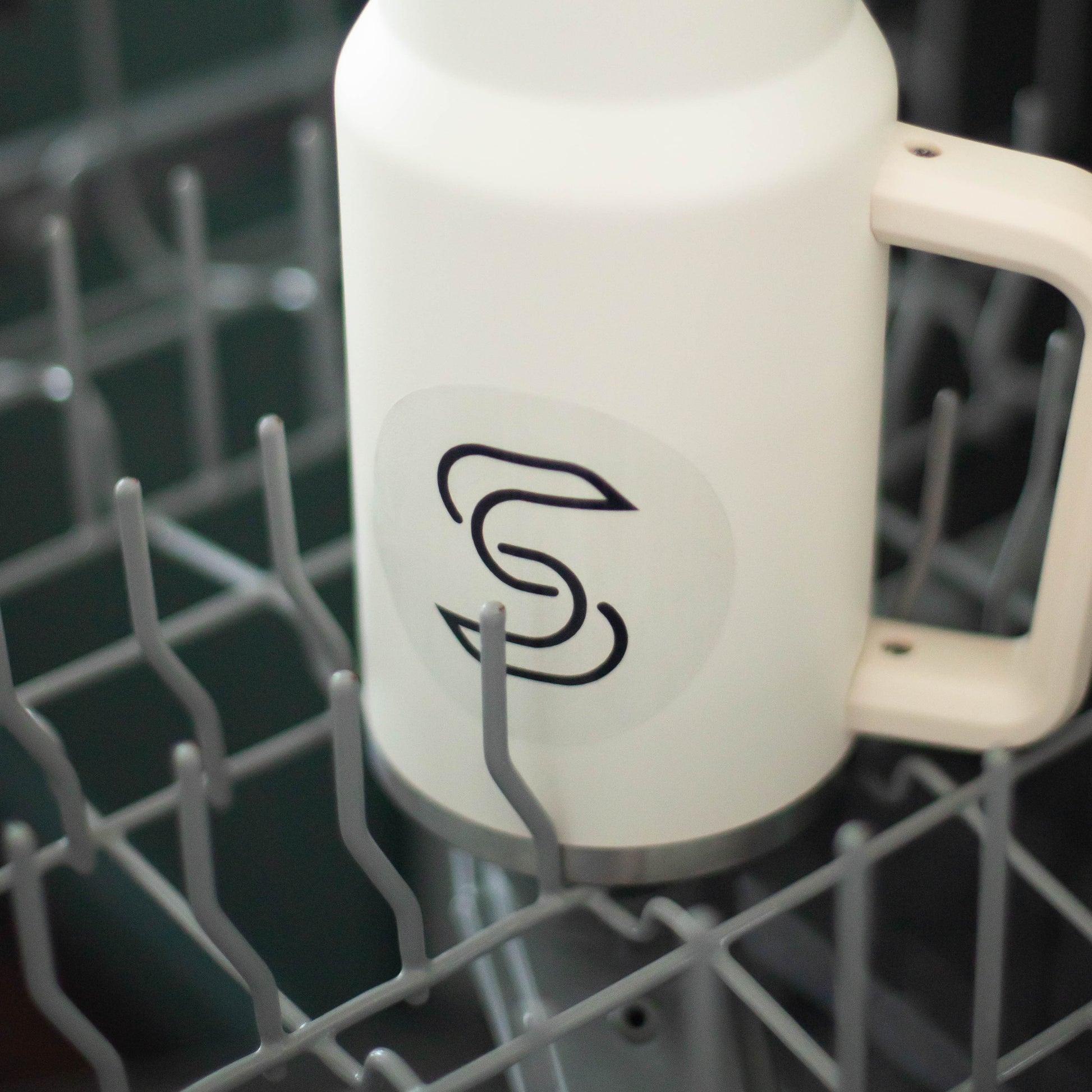 A clear circular sticker of a black Stickr logo on a water bottle in a dishwasher