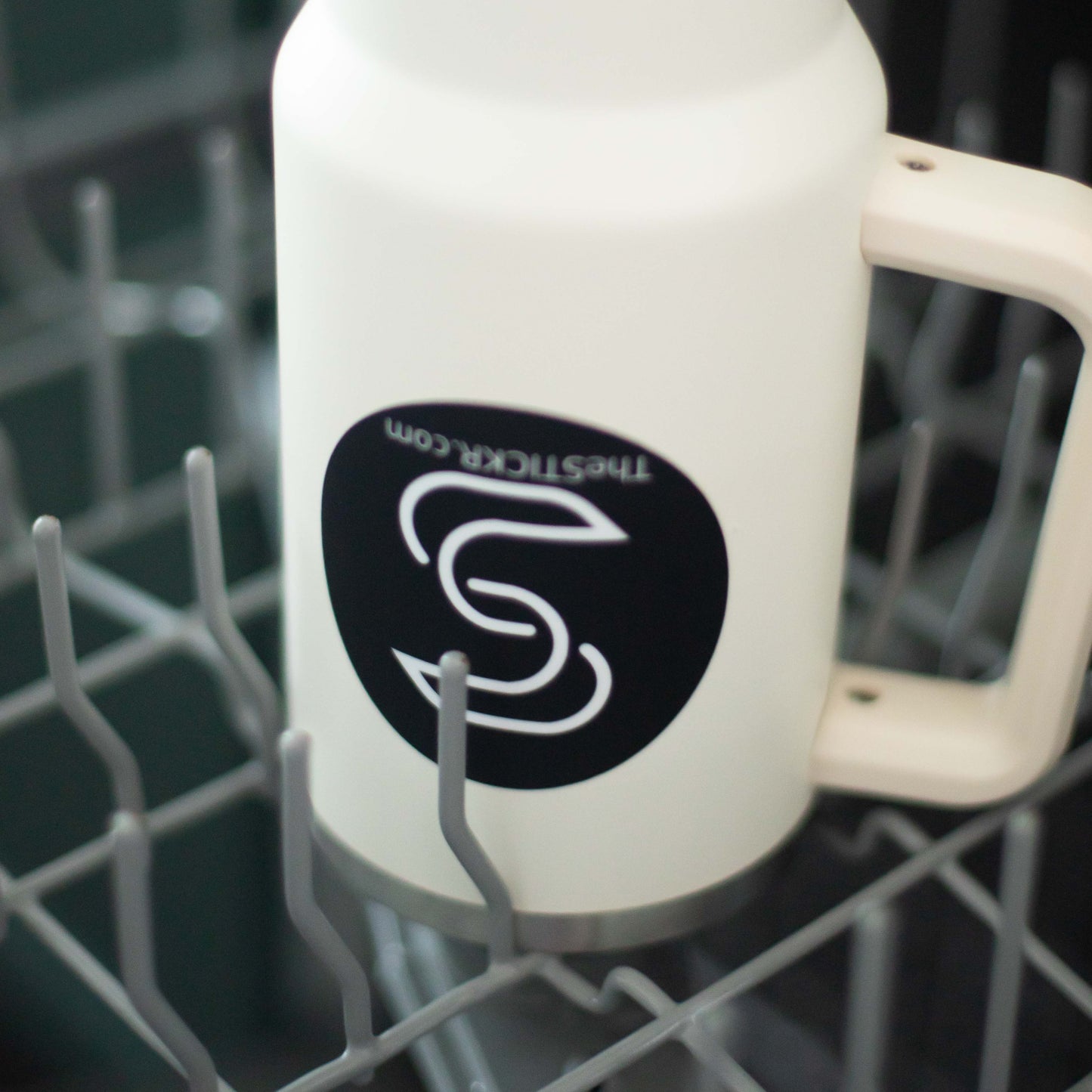 A black circular vinyl sticker of the Stickr logo and website on a water bottle in a dishwasher