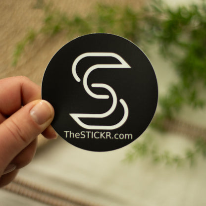 A hand holding a black circular vinyl sticker of the Stickr logo and website