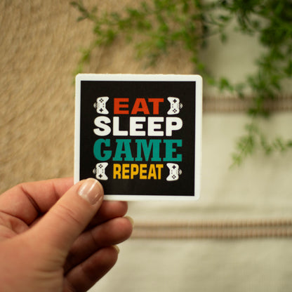 A hand holding a square-shaped sticker with the words eat sleep game repeat and images of game controllers