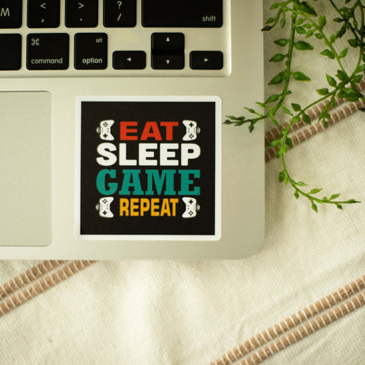 A square-shaped sticker with the words eat sleep game repeat and images of game controllers on a laptop