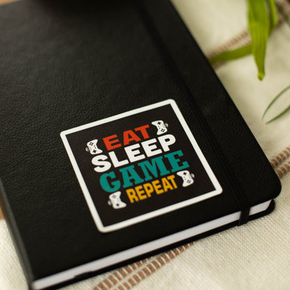 A square-shaped sticker with the words eat sleep game repeat and images of game controllers on a notebook