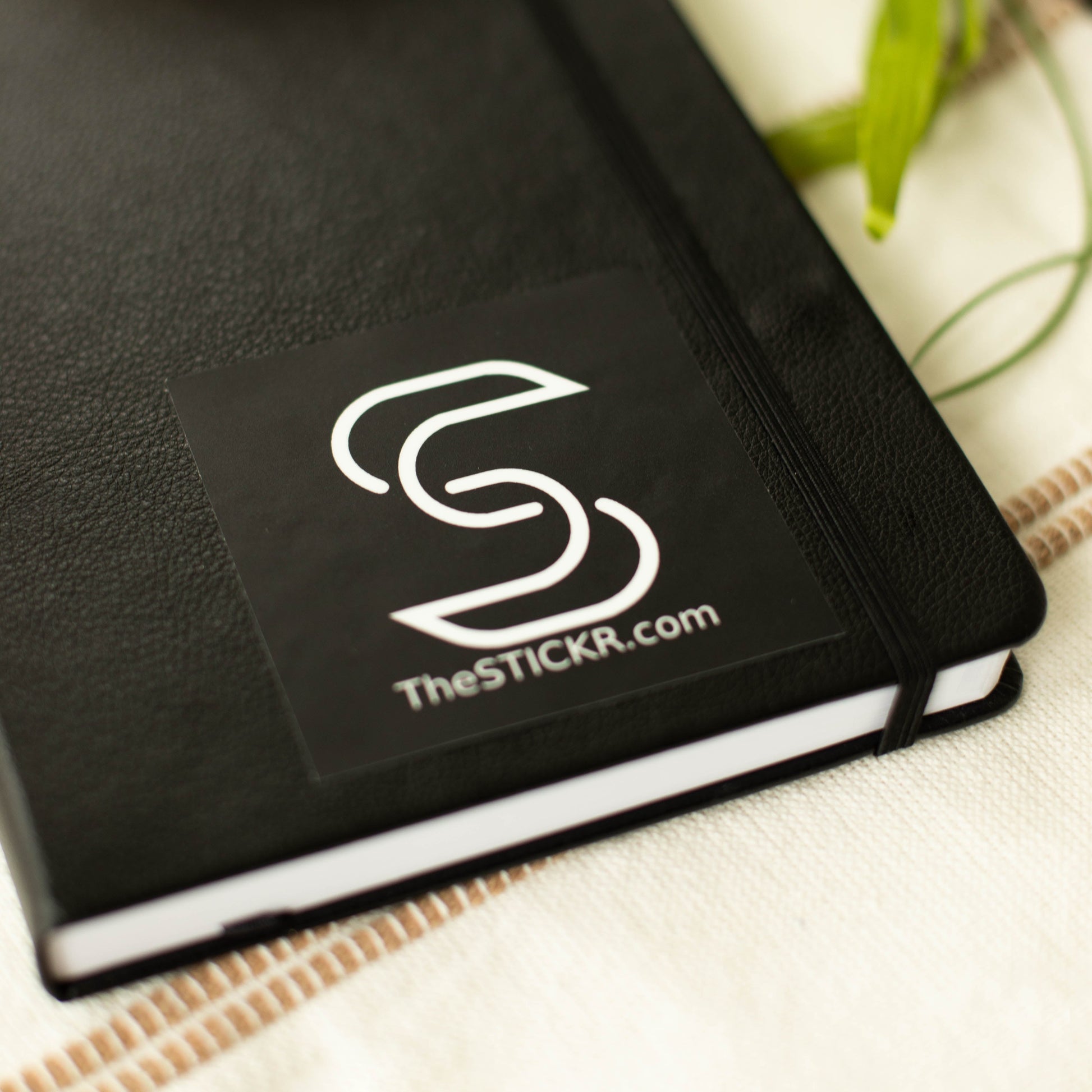 A square-shaped black vinyl sticker of the Stickr logo and website on a notebook