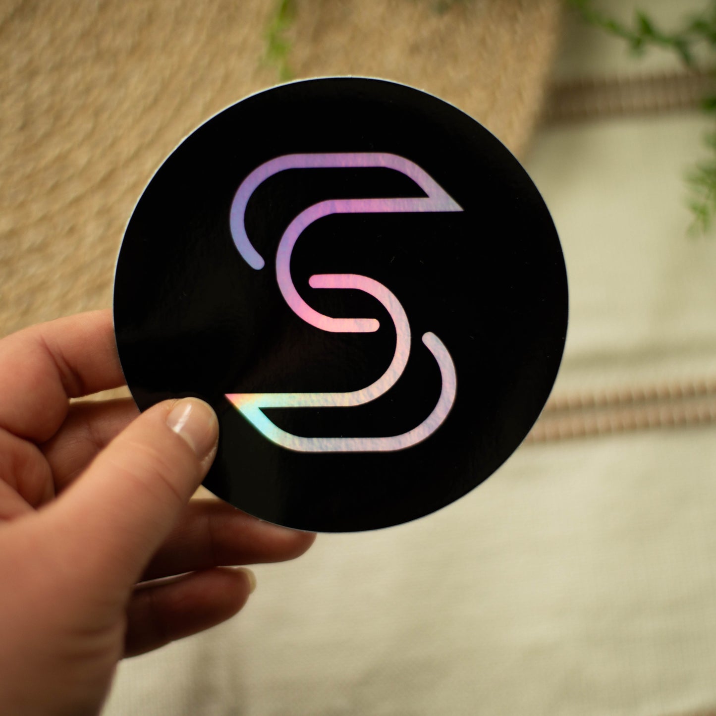 A hand holding a circular holographic sticker of the Stickr logo